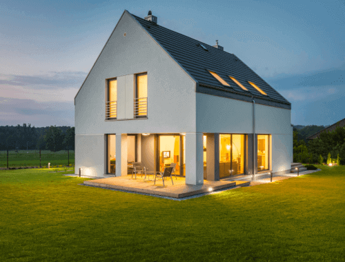 A detached house in the country side with orange lights glimmering through all of the house windows.