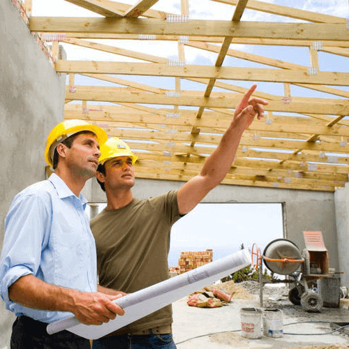 Two builders on a construction site one is holding blue prints and the other is pointing at the roof top, they both look concentrated.